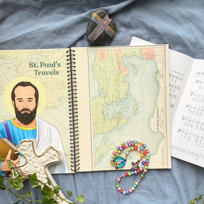 Open book titled 'St. Paul's Travels' with a map, rosary, and stone on a blue fabric background.