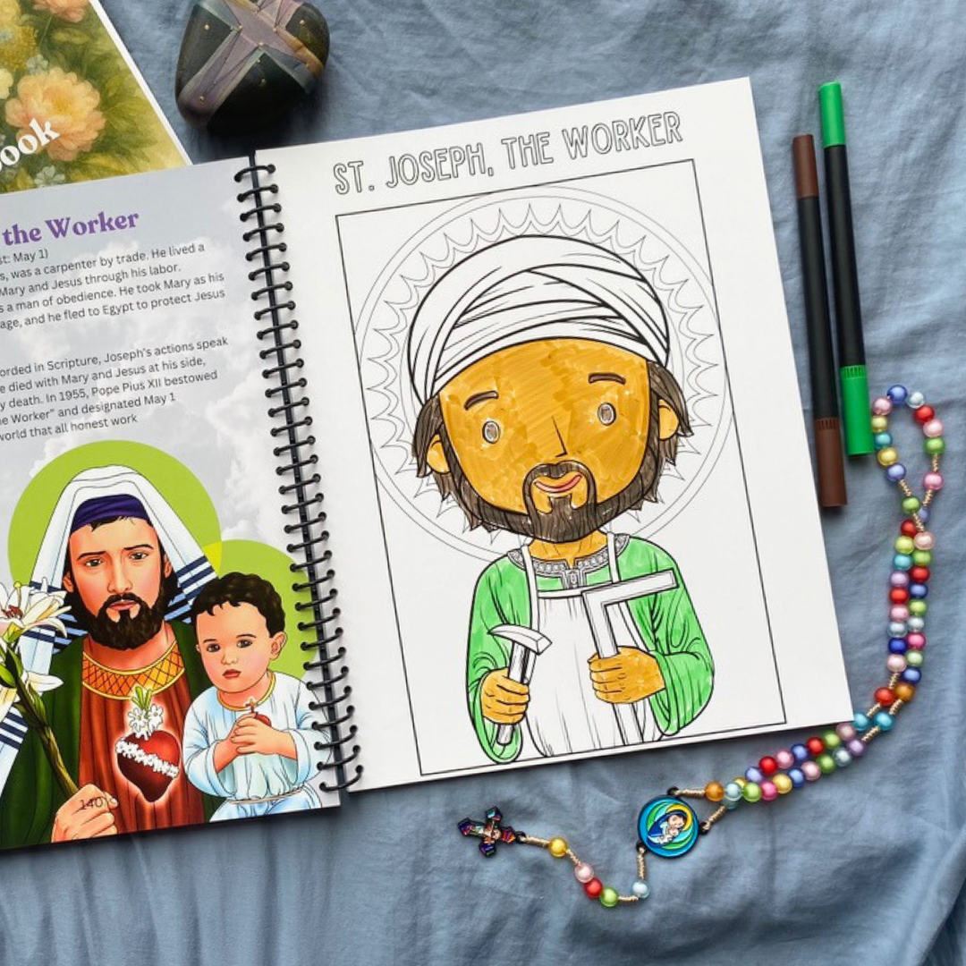 Coloring book open to a page with a illustration of St. Joseph the Worker, surrounded by coloring supplies on a blue fabric background.